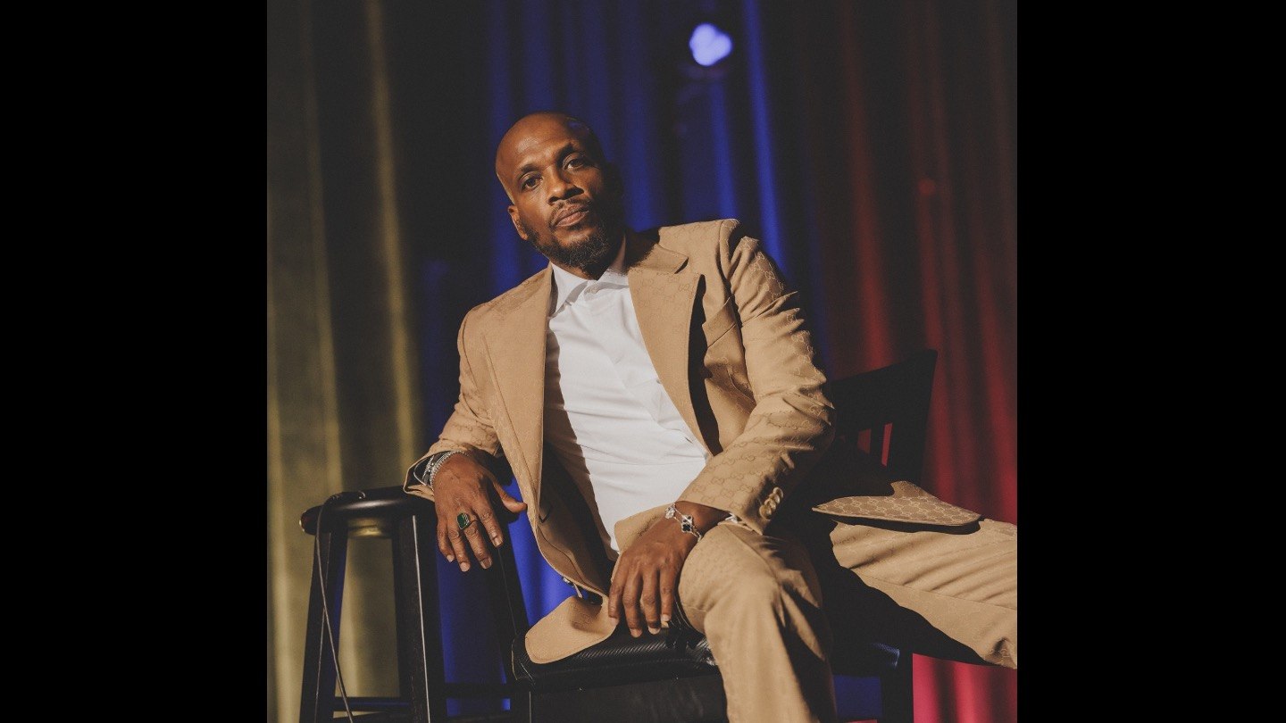 Comedian Ali Siddiq in a tan suit, seated on a stool in front of blue and red stage curtains