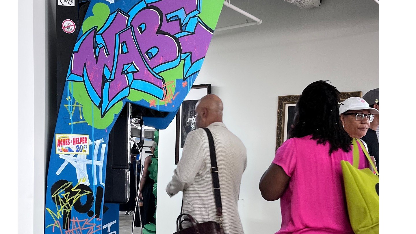 A tall booth painted with colorful WABE graffiti lettering stands in a white gallery as visitors walk past framed art