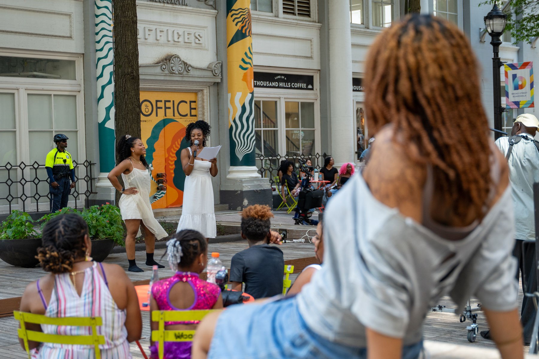 Two performers in white dresses, one reading into a microphone, perform on the Broad Street Boardwalk as an audience watches from yellow cafe chairs in front of a historic white building.