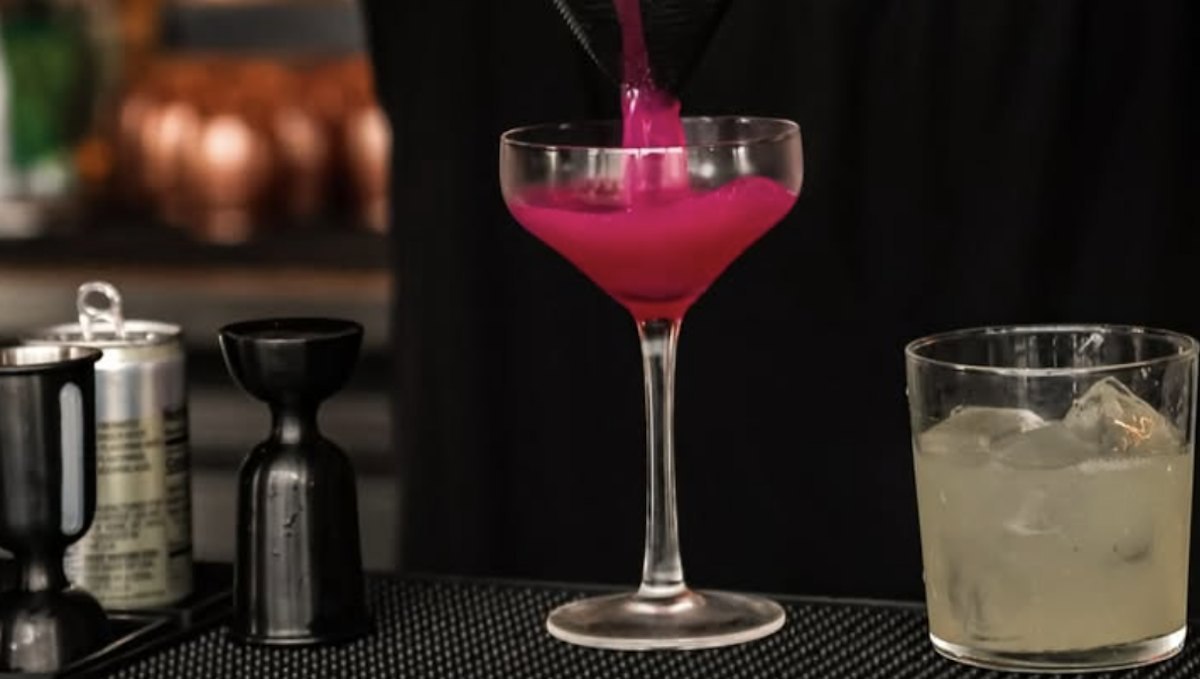 A bright pink cocktail being poured into a coupe glass on a bar top beside a jigger and a glass of iced drink