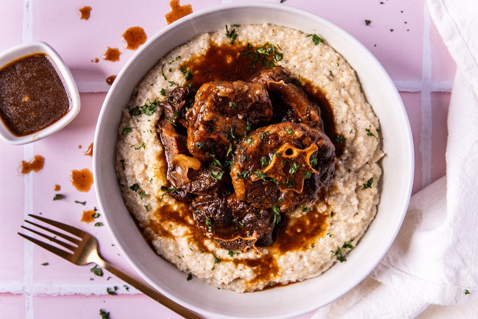 A white bowl of creamy stone ground grits topped with saucy oxtail with fork beside the bowl on a pink background from Betty Sue's in Atlanta.