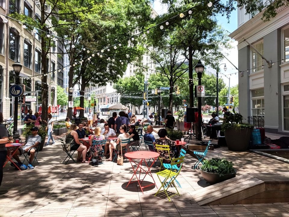People seated at colorful bistro tables under string lights and shade trees on the Broad Street Boardwalk plaza in downtown Atlanta's Fairlie-Poplar District