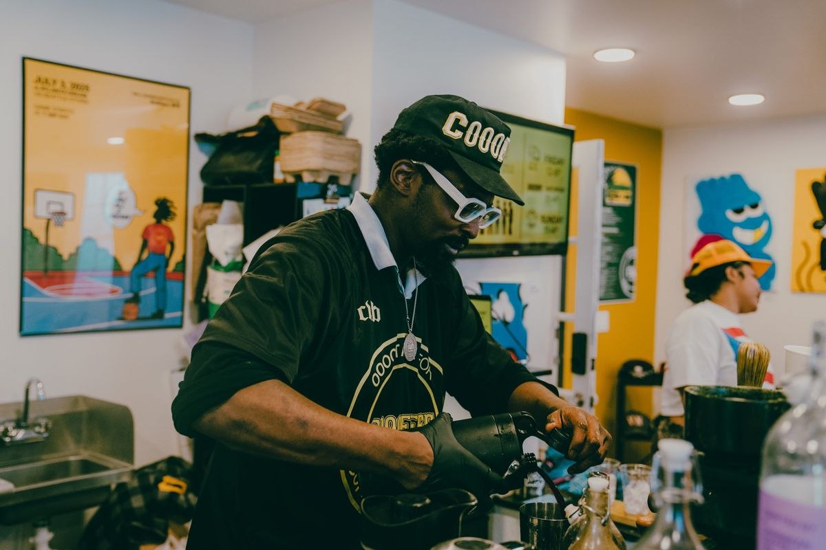 Coffee Was Black owner Malcolm Voltaire, in a black cap and white glasses, preparing a drink behind the counter with colorful artwork on the walls behind him