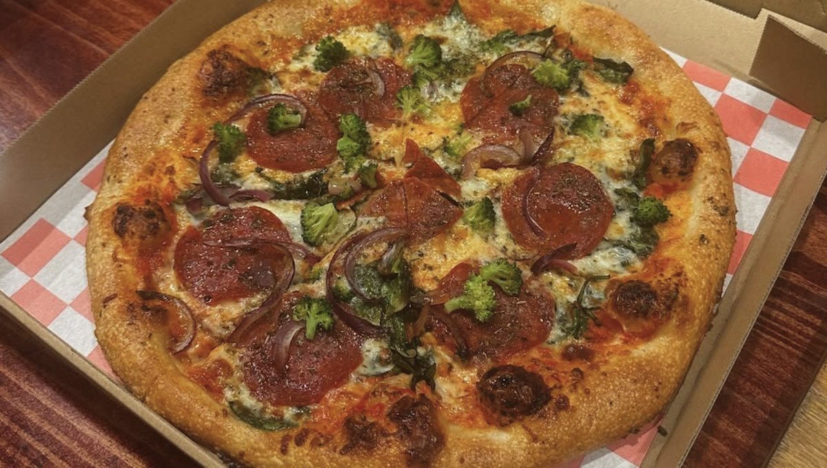 Dolo's Pizza pie topped with pepperoni, broccoli, red onion and greens in a takeout box on red-checked paper