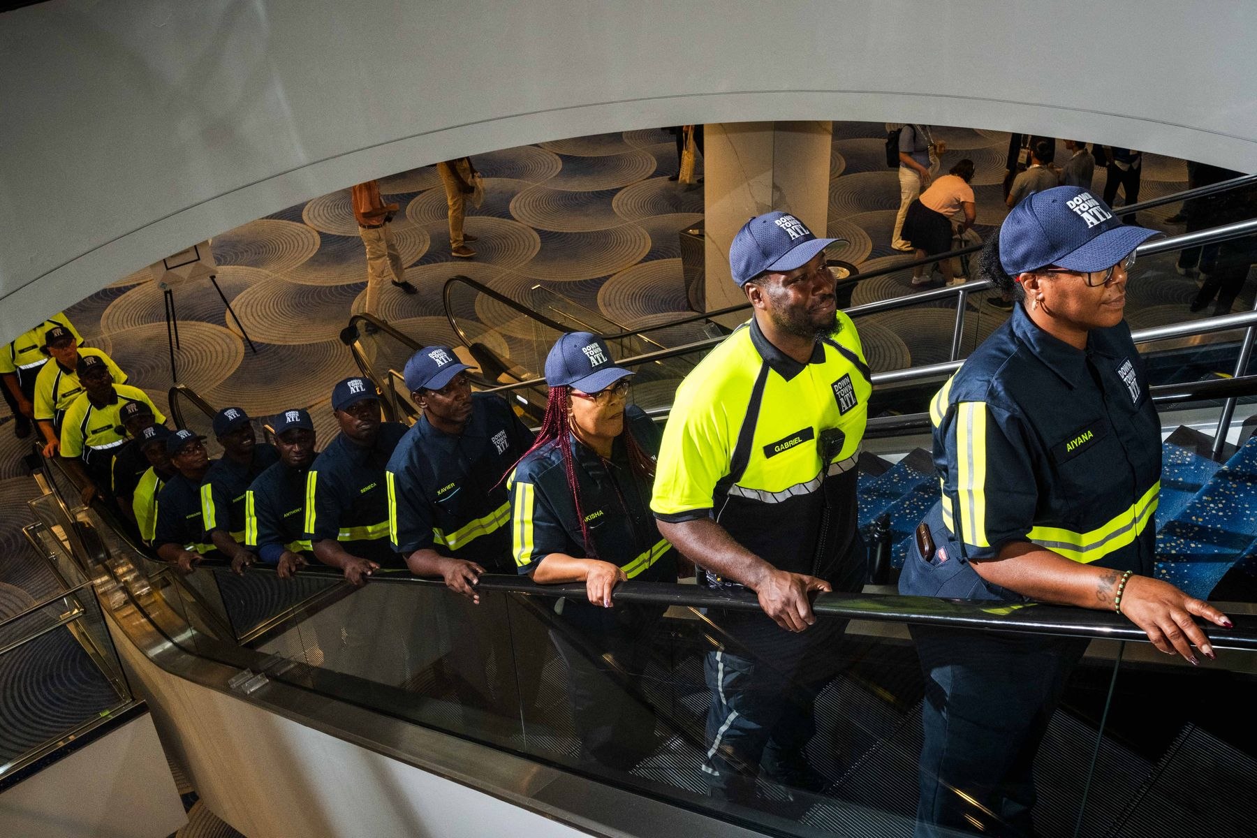 Downtown Ambassadors in new navy and bright yellow uniforms and caps ride an escalator in a hotel atrium