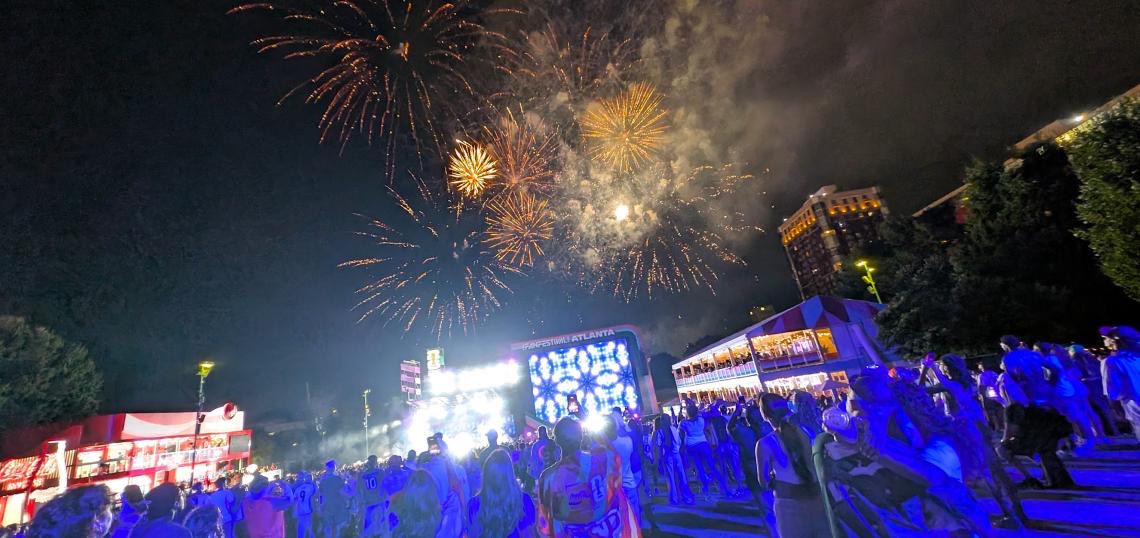 Fireworks burst over the FIFA Fan Festival stage at Centennial Olympic Park at night as a crowd bathed in blue light watches.