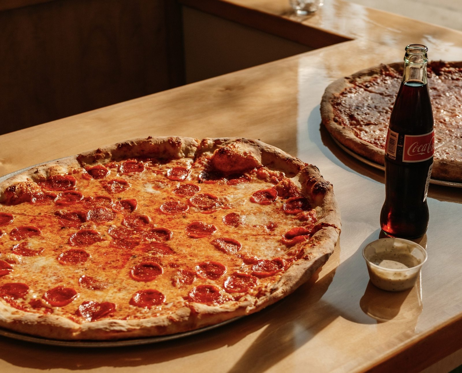 A whole pepperoni pizza and a whole cheese pizza on a wooden counter beside a glass bottle of Coca-Cola at sunset.