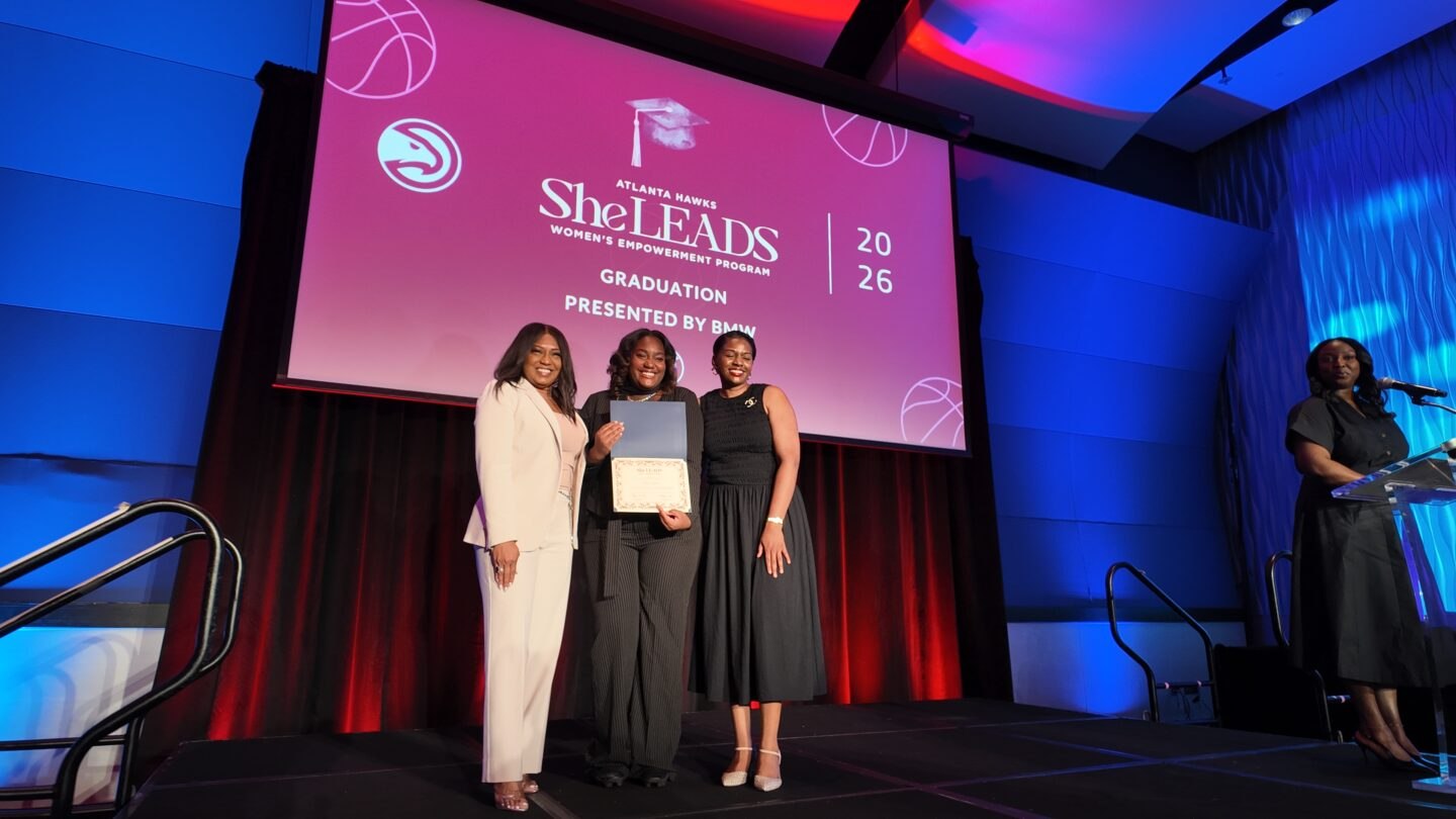 Three women pose on stage with a graduation certificate in front of a screen reading Atlanta Hawks She LEADS 2026 Graduation, while a speaker stands at a podium