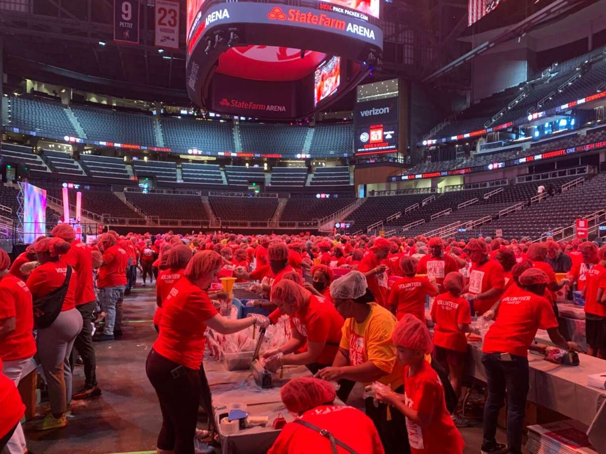 Hundreds of volunteers in red T-shirts and hairnets pack meals at long tables on the State Farm Arena floor