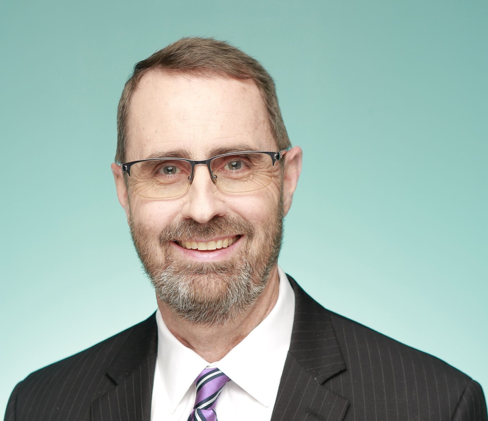 Kevin Duvall, new CEO of the Georgia World Congress Center Authority, smiling in glasses, a dark pinstripe suit and a purple striped tie against a teal backdrop