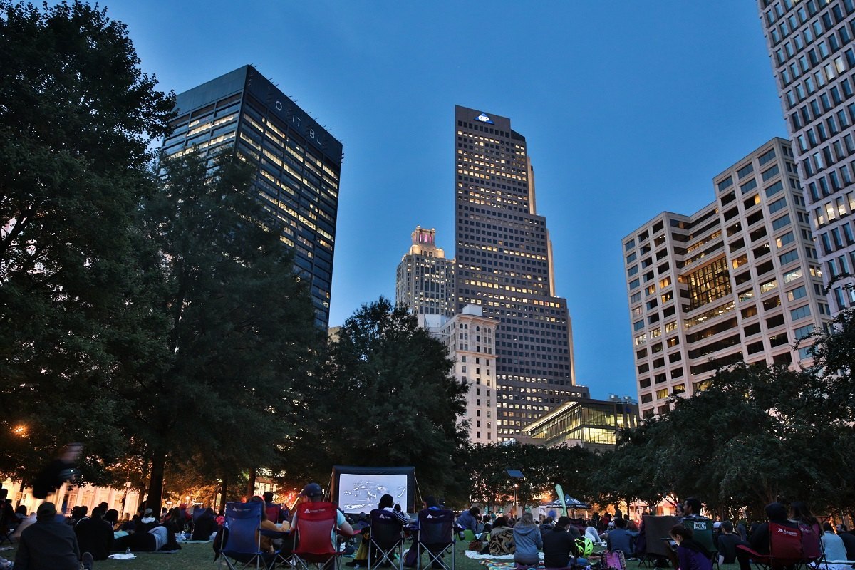 Crowd in lawn chairs and on blankets at dusk in Woodruff Park, with lit downtown Atlanta towers rising behind the trees