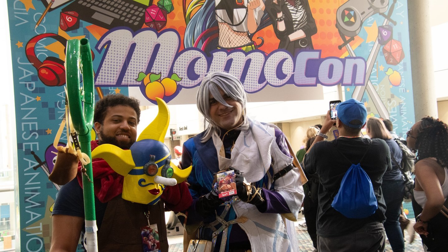 Two costumed fans pose in front of a large colorful MomoCon banner while other attendees take photos inside the convention center