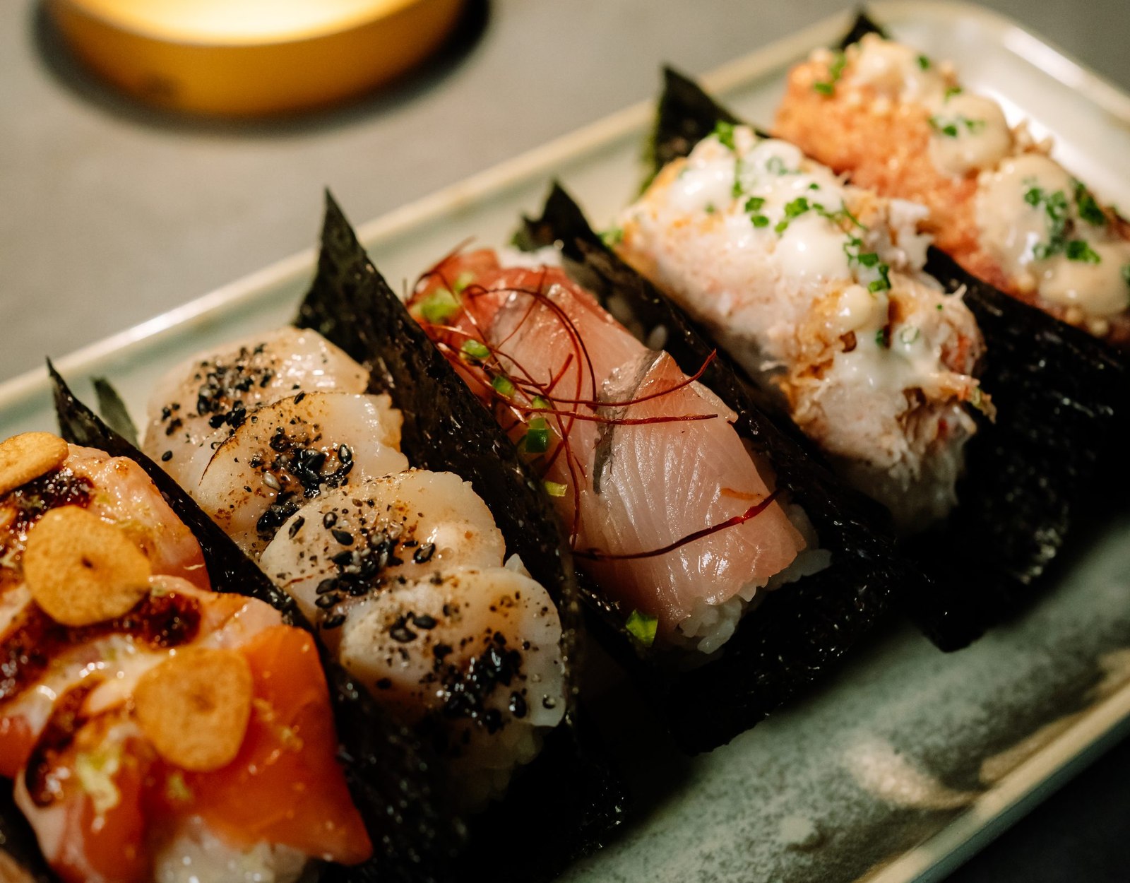 A row of NoriFish hand rolls topped with scallop, yellowtail and crab on a ceramic plate