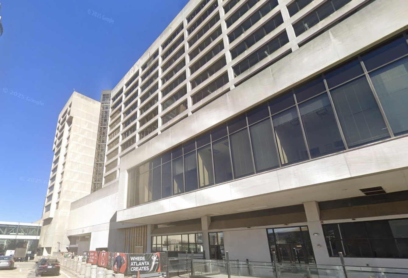 Street-level view of the concrete and glass facade of the former CNN Center building in downtown Atlanta under a clear blue sky, with a Where Atlanta Creates banner along the sidewalk