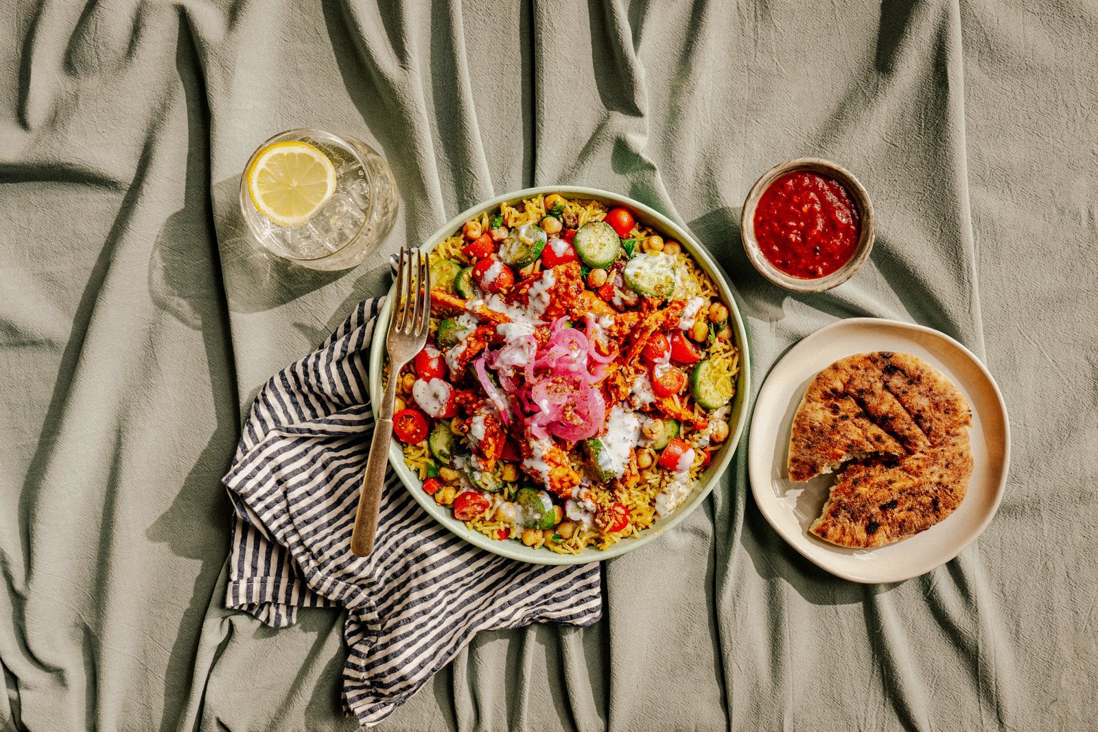 Overhead view of a Roti rice bowl with chicken, chickpeas, cucumbers, tomatoes and pickled onions beside pita and a cup of red sauce