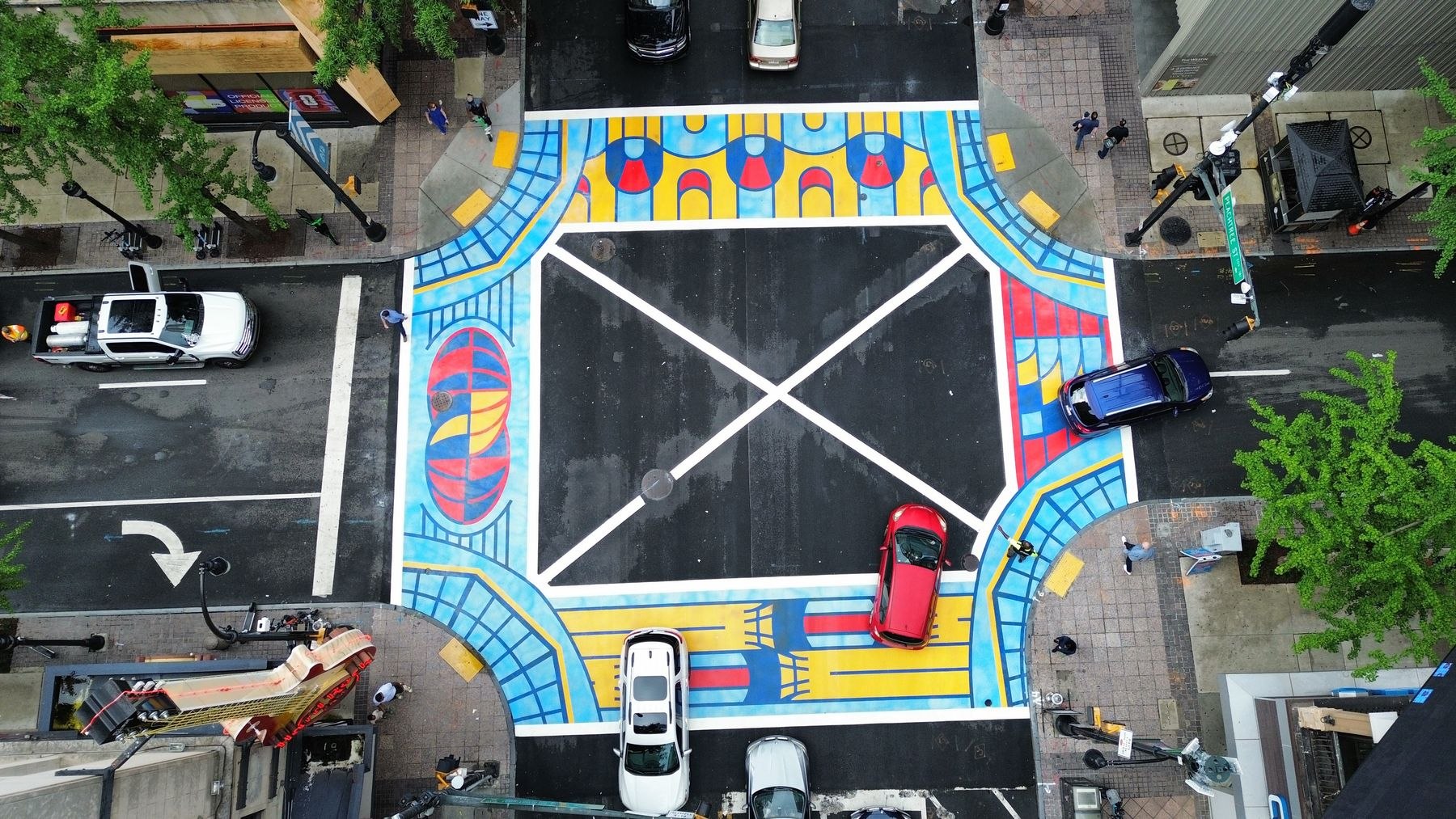 Overhead drone view of the Peachtree Street and Andrew Young International Boulevard intersection, with all four crosswalks painted in bright blue, yellow and red patterns as cars pass through.