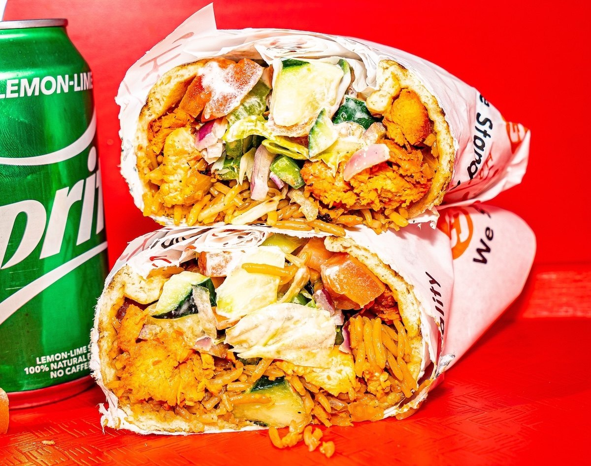 Two halves of a Shah's Halal Food chicken wrap stacked in branded paper next to a lemon-lime soda can on a red background