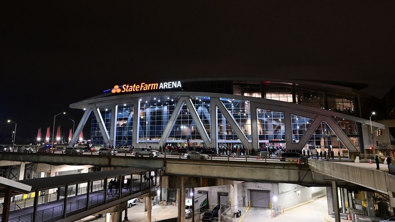 State Farm Arena in downtown Atlanta lit up at night, with crowds gathered outside the glass entrance