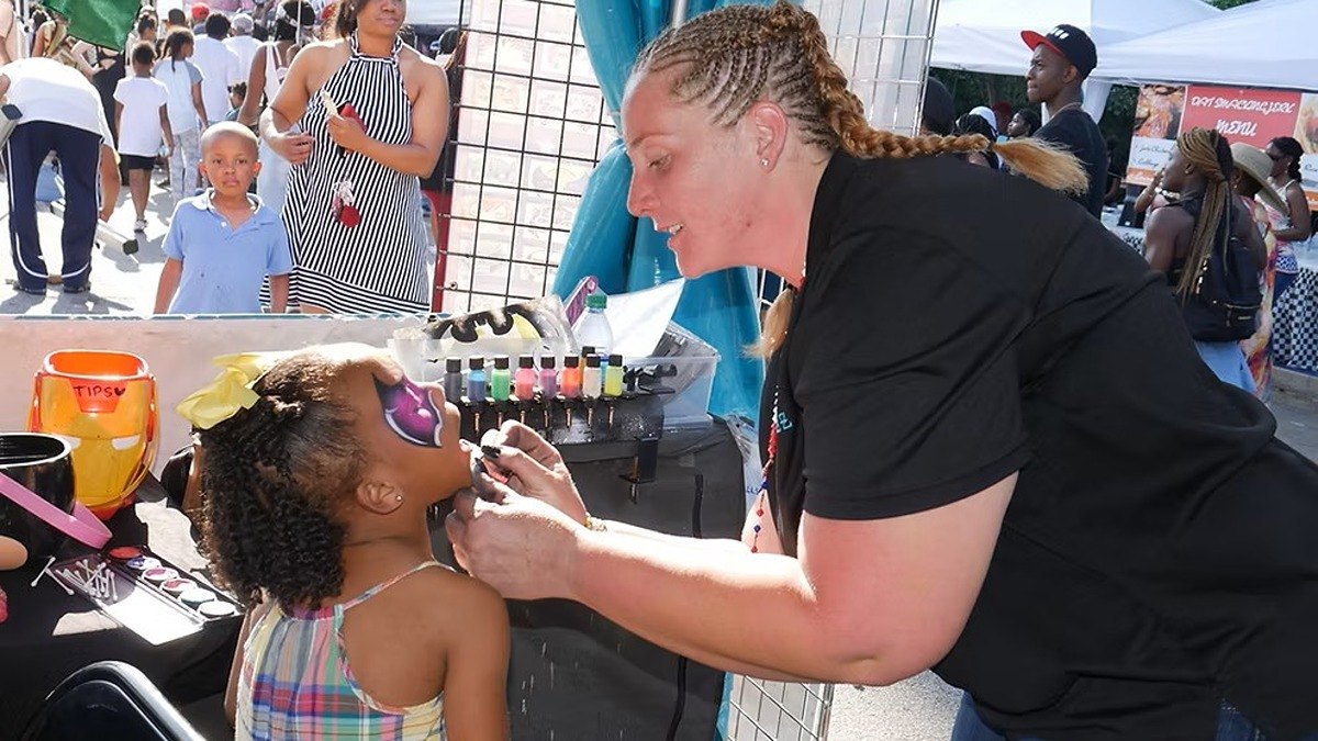 A face painter decorates a young girl's face at a vendor booth while festivalgoers walk past at Sweet Auburn Springfest