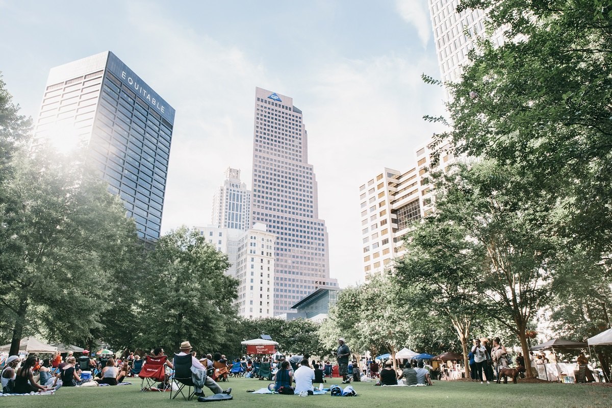 People relax on blankets and lawn chairs on the grass at Woodruff Park, with the Equitable Building and Georgia-Pacific tower rising above the trees