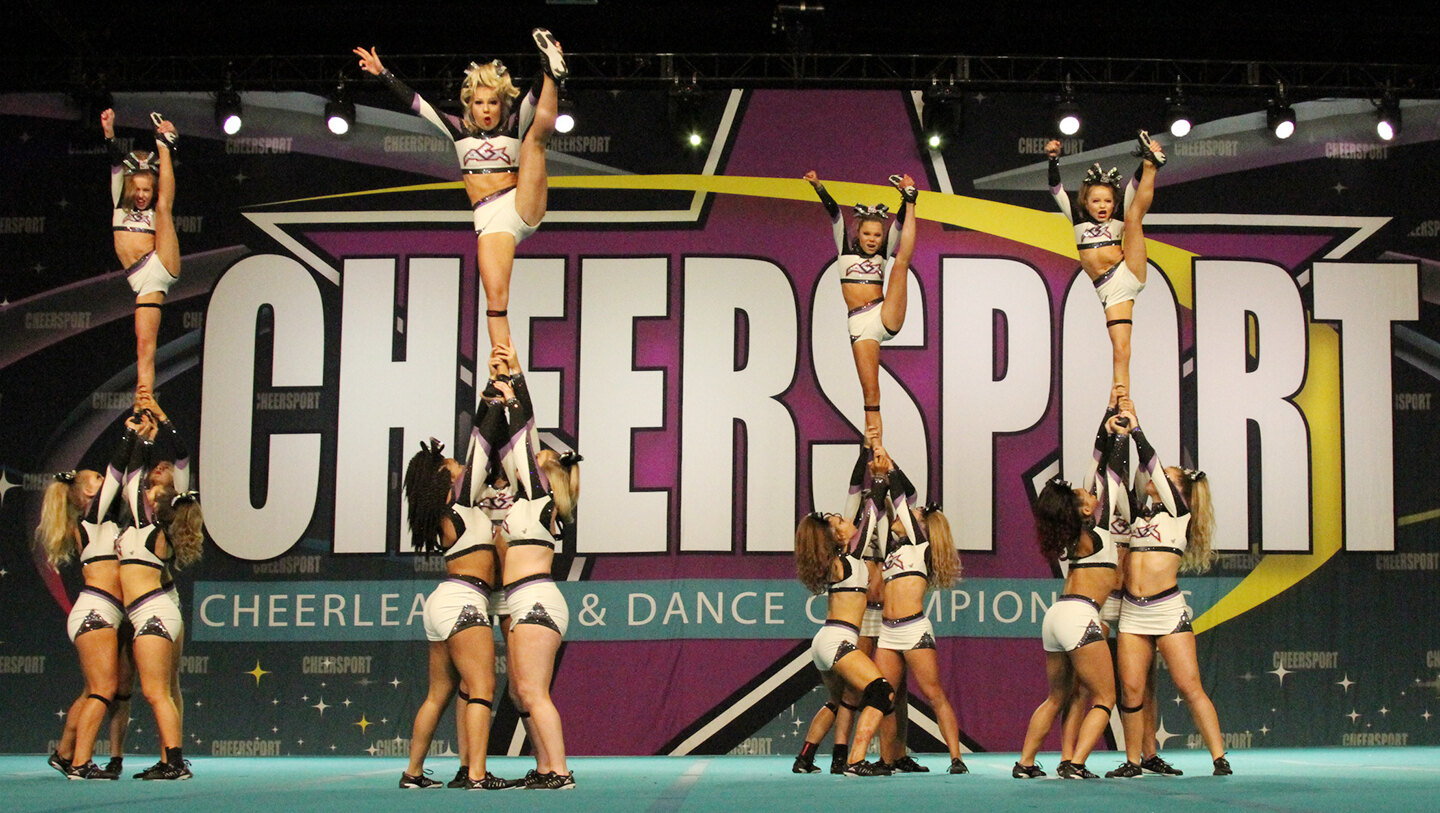 All-star cheerleaders in white and purple uniforms hold four extended stunt groups with flyers in vertical splits on a teal mat before a giant CHEERSPORT Cheerleading and Dance Championship backdrop