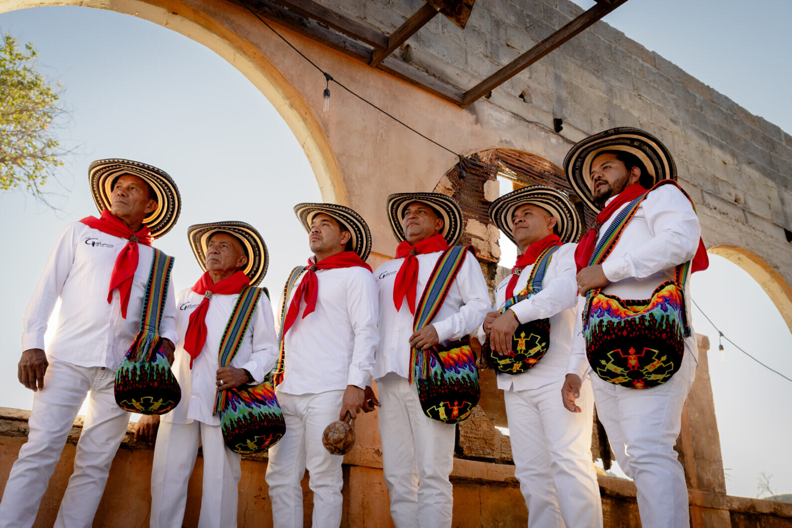 Los Gaiteros de San Jacinto — six musicians in white outfits, red neckerchiefs and sombrero vueltiao hats with woven mochila bags, beneath a crumbling arched ruin — Madre Cumbia at The Masquerade