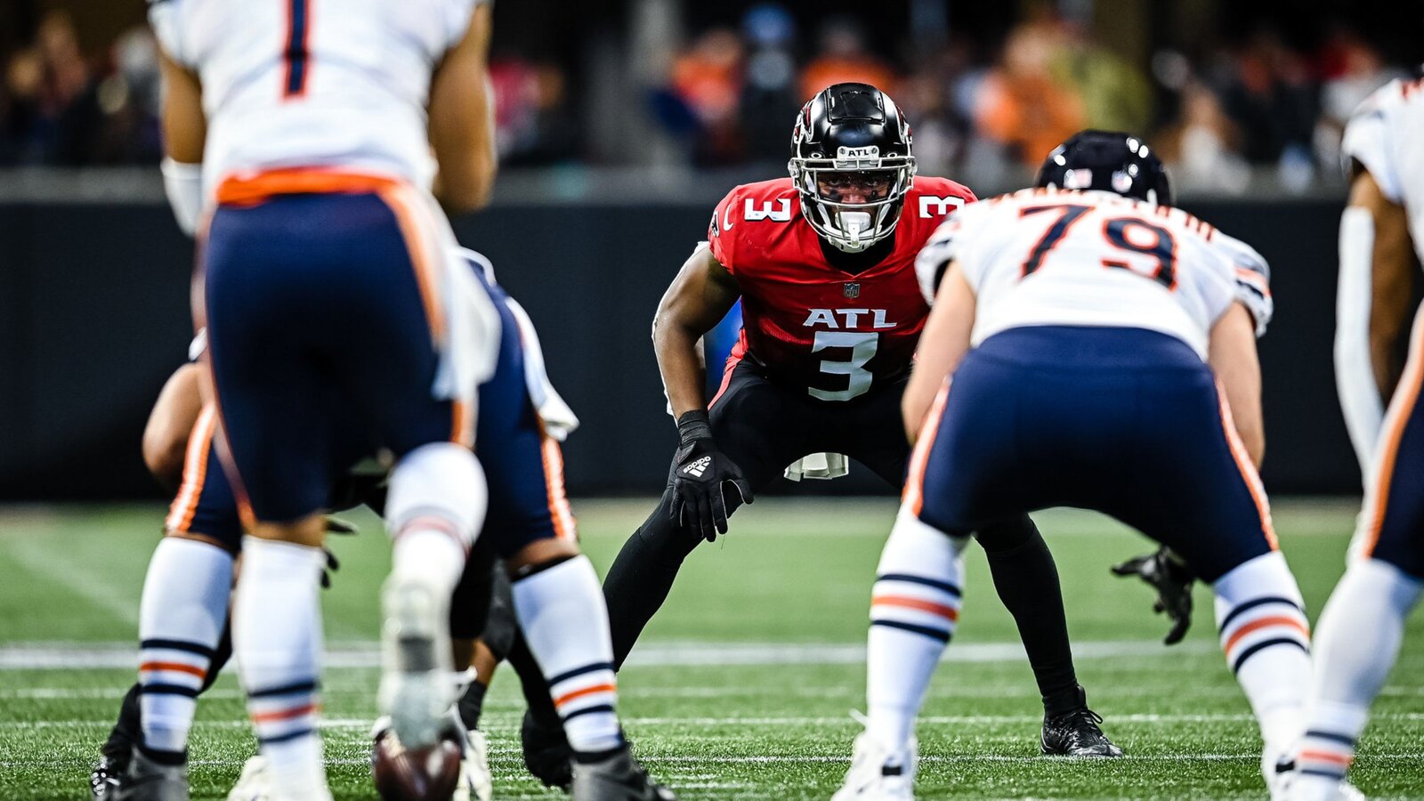 An Atlanta Falcons defender in a red jersey and black ATL helmet crouches in a stance on the turf at Mercedes-Benz Stadium, facing Chicago Bears offensive linemen in white and navy road uniforms.