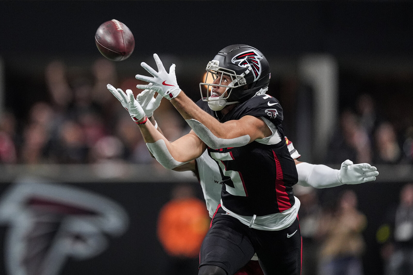 An Atlanta Falcons wide receiver in the black home uniform stretches both arms overhead to haul in a pass as a defender trails him at Mercedes-Benz Stadium