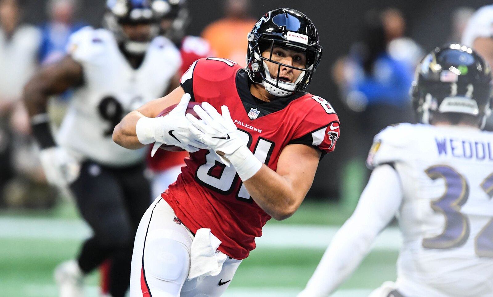 Atlanta Falcons tight end in a red home jersey tucks the football and runs upfield as Baltimore Ravens defenders in white road jerseys close in, on the turf at Mercedes-Benz Stadium.