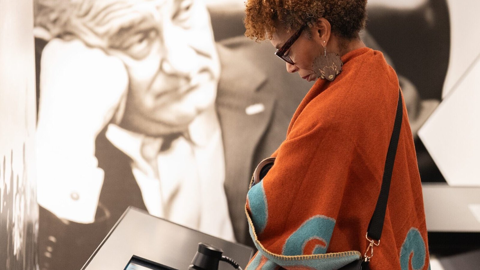 A visitor in an orange patterned shawl leans over an interactive touchscreen exhibit at the National Center for Civil and Human Rights, in front of a large black-and-white mural of President Lyndon B. Johnson