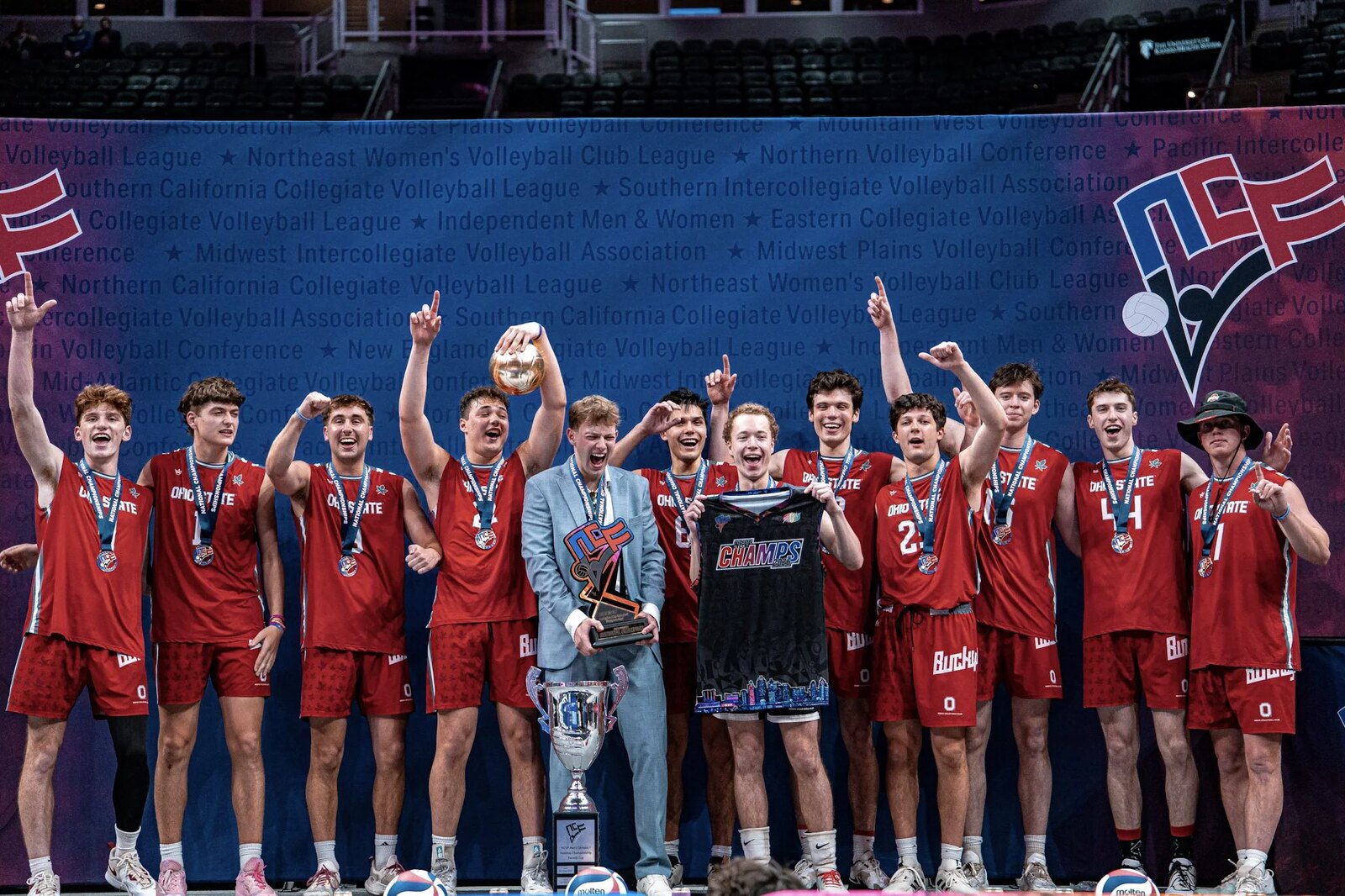 A mens club volleyball team in red uniforms celebrates on the podium with the NCVF championship trophy and medals in front of the NCVF member-league backdrop