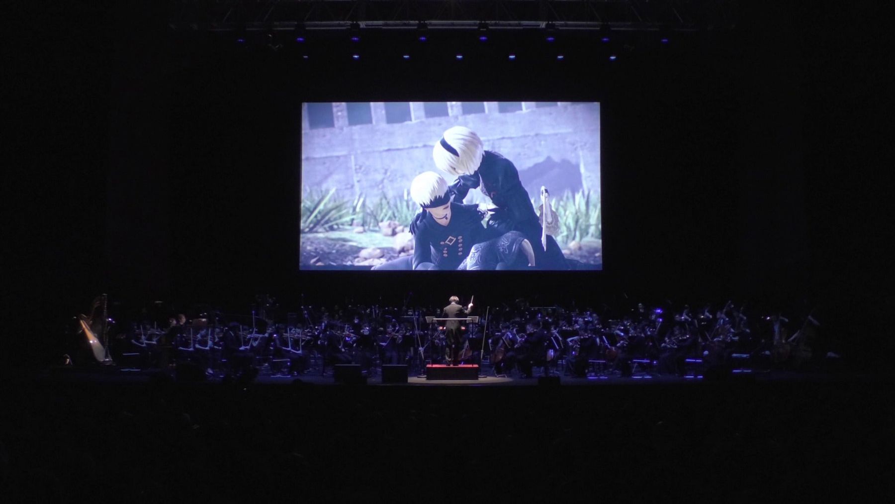 A full orchestra performs beneath a giant screen showing NieR game footage