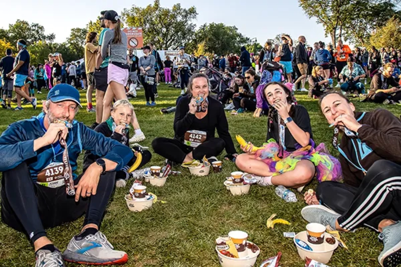 Runners in a park after a Hot Chocolate Run, biting their finisher medals and holding post-race snacks
