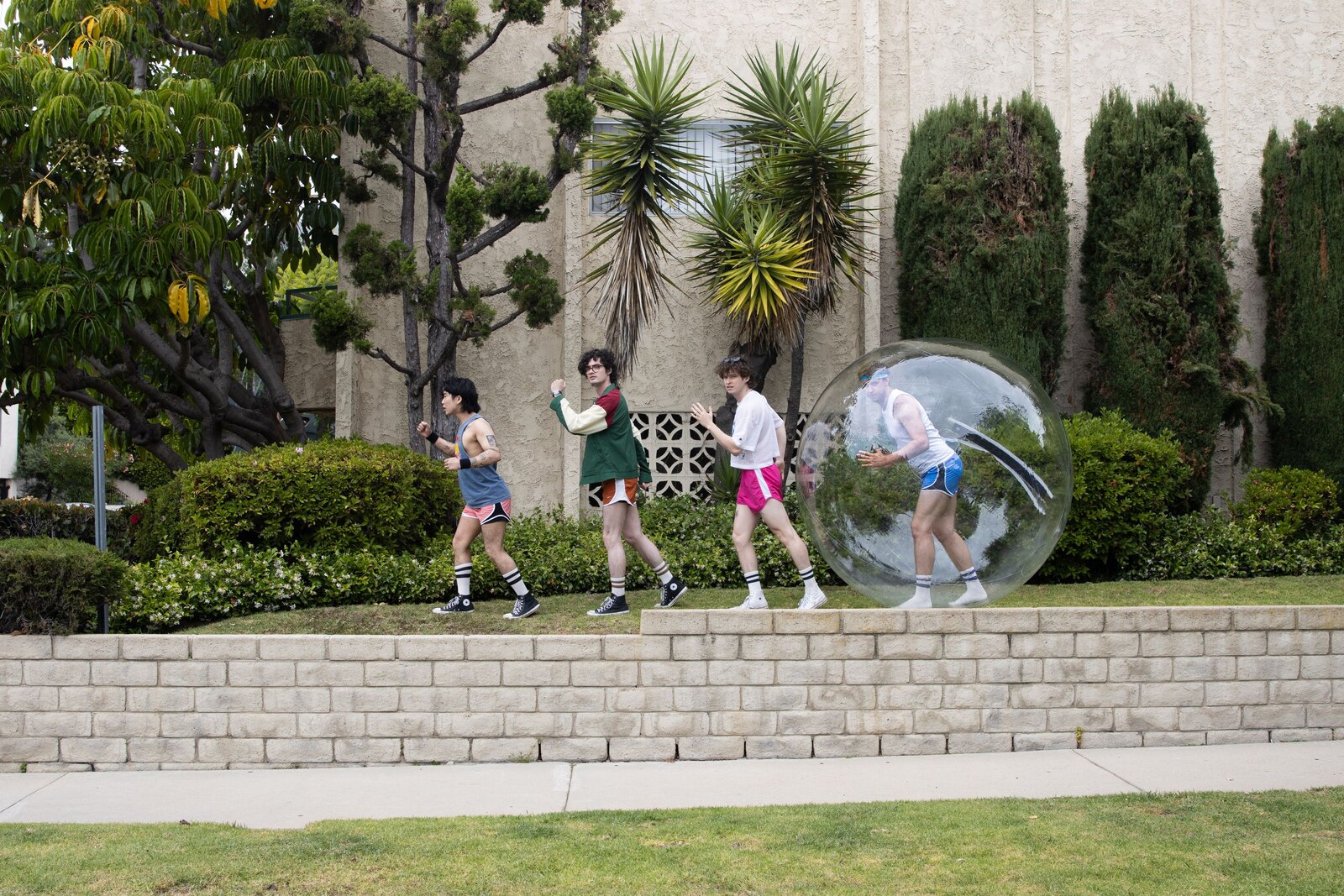Sorry Ghost, the four-piece Los Angeles indie rock band, in retro running gear jogging single-file along a low brick wall past palms and hedges, the last member sealed inside a giant clear plastic bubble