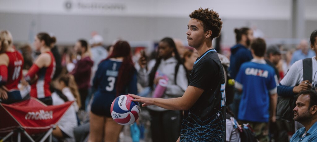 A young volleyball player in a black and teal jersey holds a Molten volleyball on a crowded convention-hall tournament floor at the Southern Region Volleyball Association championships