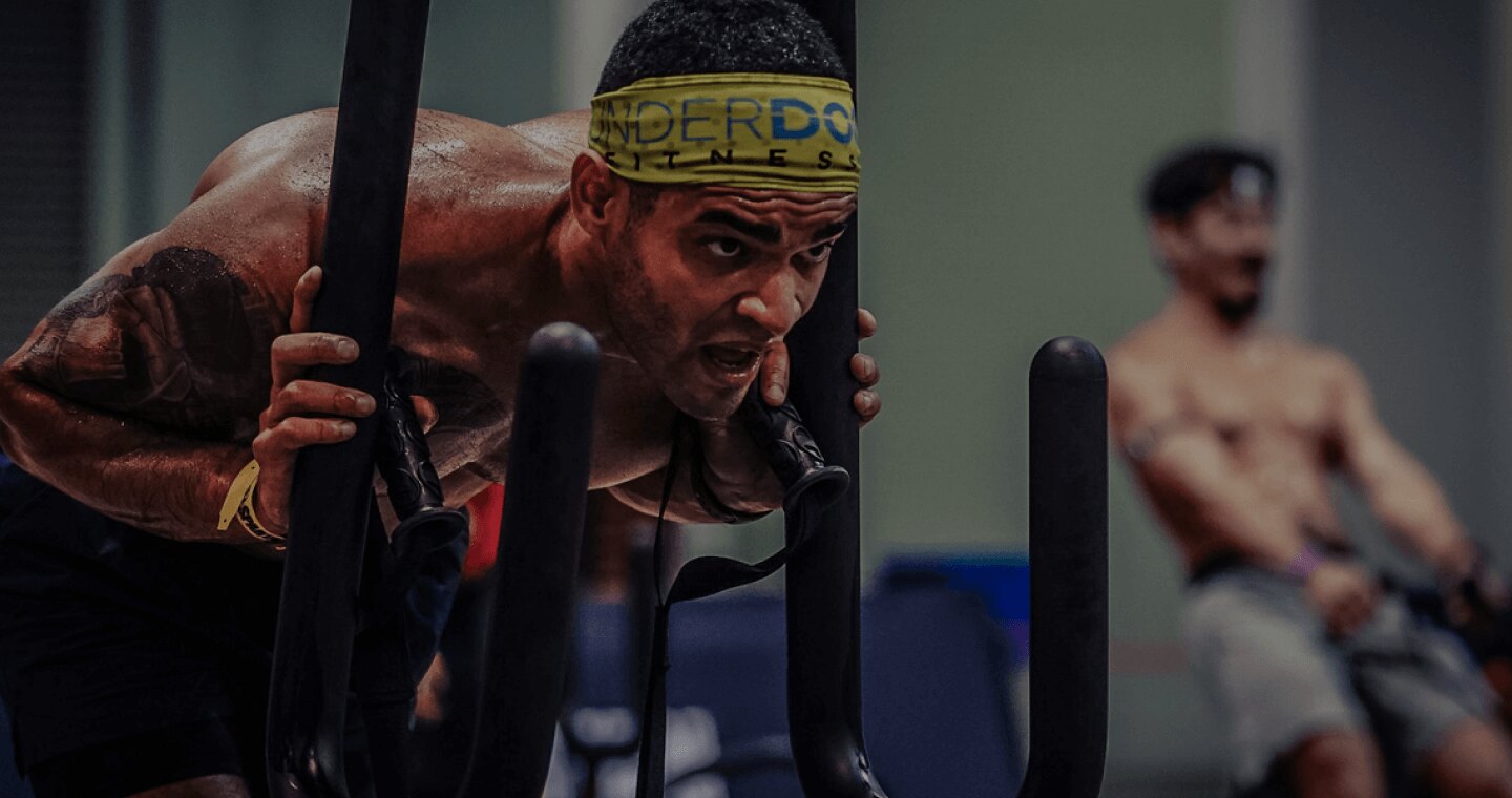 A DEKA FIT competitor in a yellow headband drives a weighted sled forward while a second athlete works a rowing machine behind him on an indoor DEKA race floor