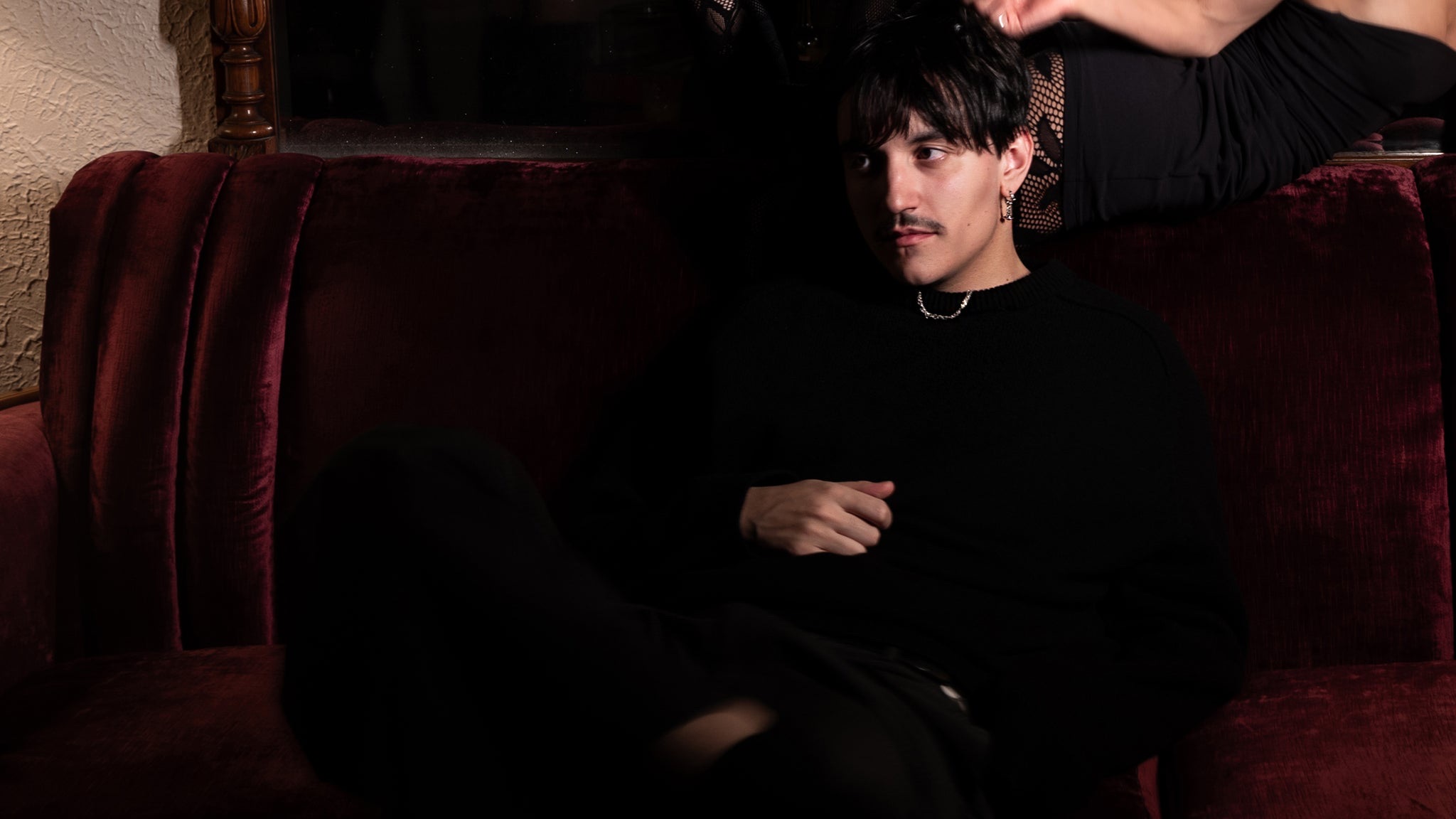 Artemas reclining on a deep red velvet couch in an all-black sweater and trousers, dark bowl-cut hair and silver chain, moody low light