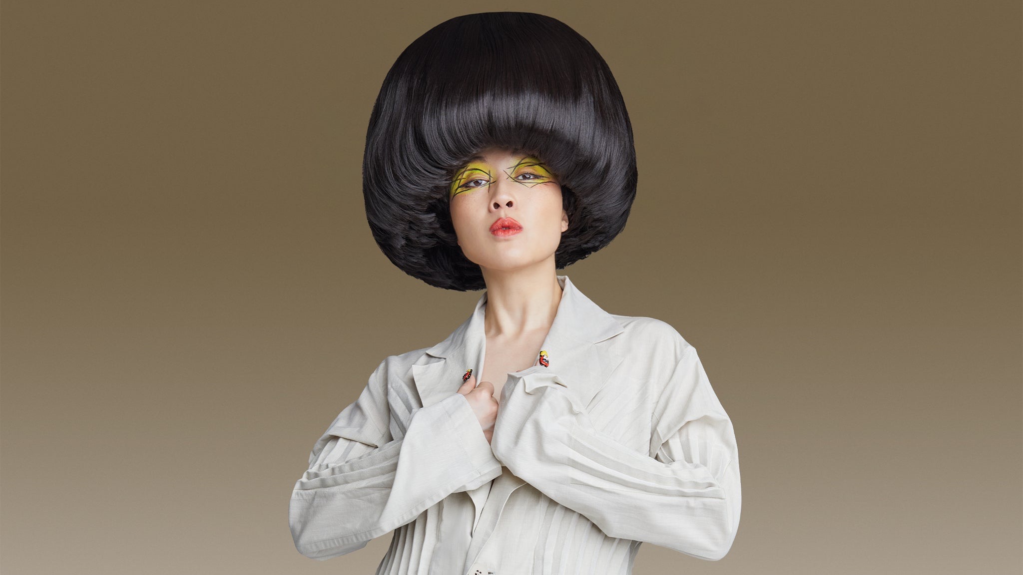 Comedian Atsuko Okatsuka in her signature oversized black bowl-cut hairstyle and yellow graphic eye makeup, wearing a cream pleated jacket against a tan backdrop — promo art for The Big Bowl Tour.