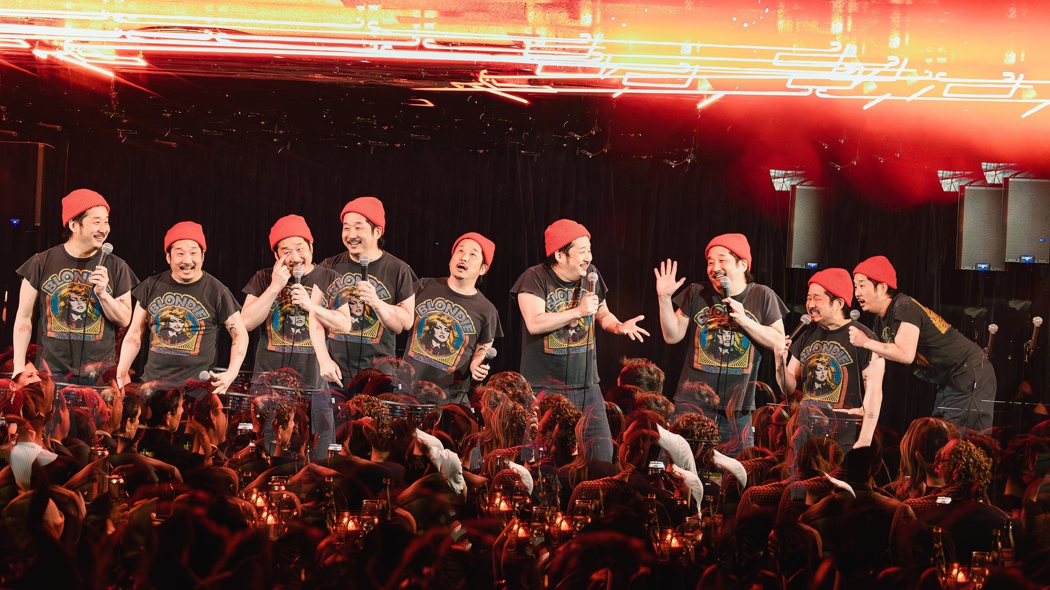 Comedian Bobby Lee performing stand-up in a multi-exposure composite, repeated nine times across a club stage in a red beanie and Blondie T-shirt holding a microphone above a red-lit crowd.