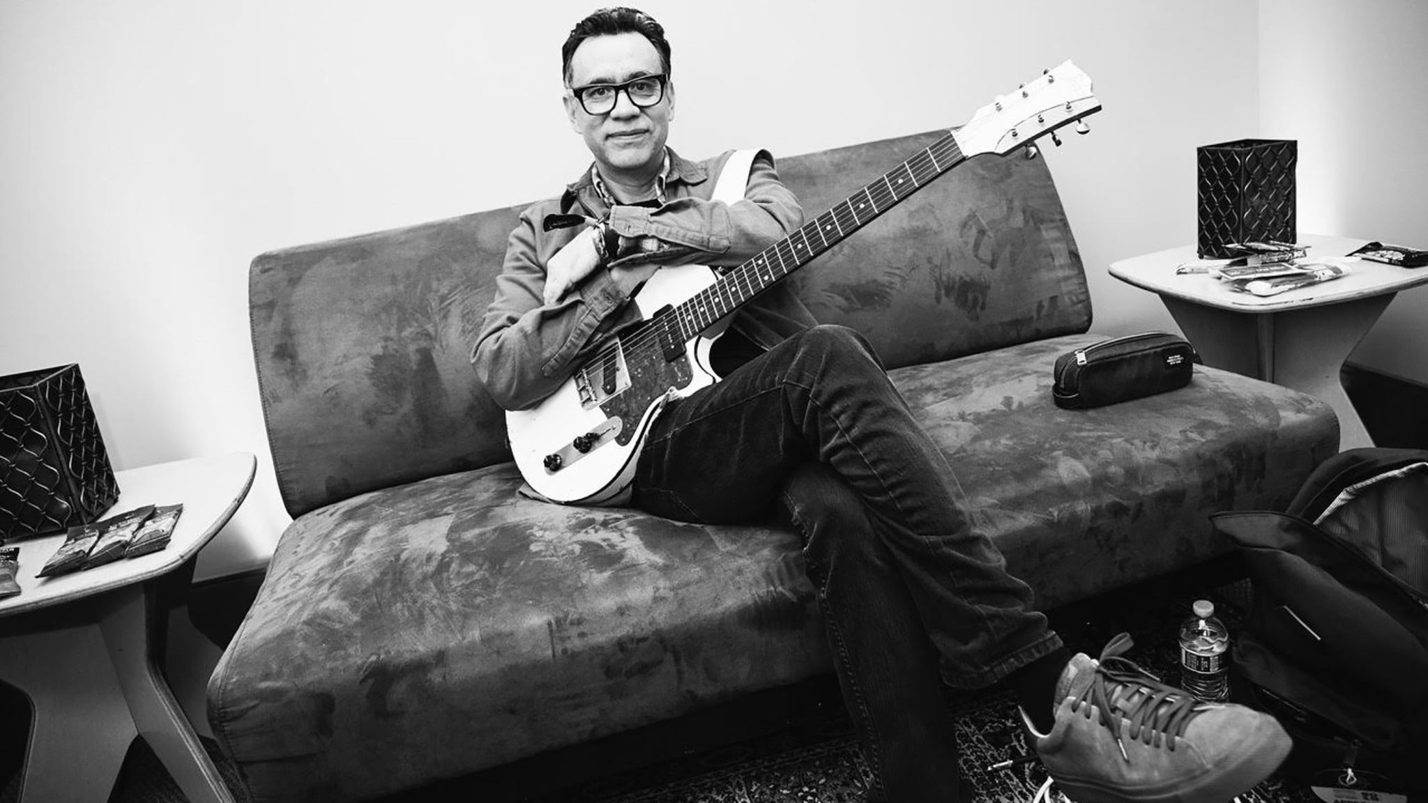 Fred Armisen in black and white, seated on a worn couch in a backstage green room, holding a white electric guitar and wearing glasses and a denim jacket.
