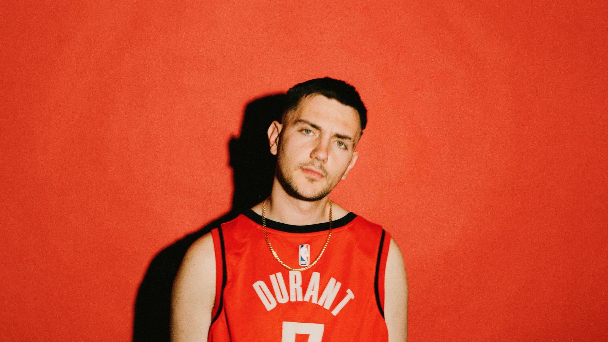 Hip-hop artist Hulvey photographed against a bright red-orange wall wearing a red Durant basketball jersey and a gold chain, from his COULD BE TONIGHT tour press kit.