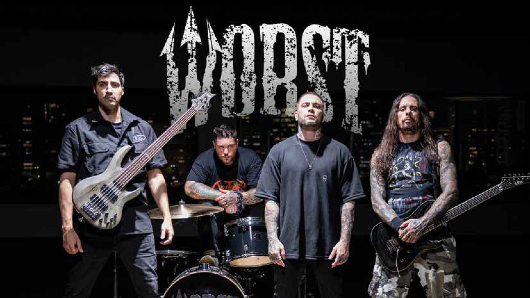 The four members of metal band Worst standing on a dark stage with bass, guitar and drum kit, the band's jagged white logo above them against a night skyline backdrop.